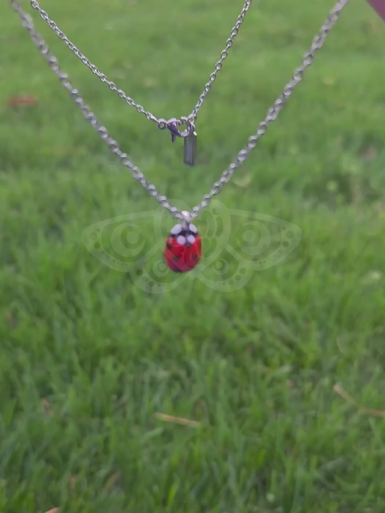 Bright red glossy ladybug pendant on dainty silver chain being handled and moved around to show details,  grass in the background.